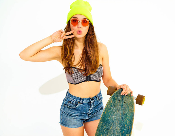 Young stylish smiling girl model in casual summer swimwear clothes and yellow beanie posing with longboard desk, showing peace sign. Isolated on white - Foto, afbeelding
