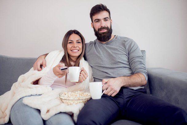 Onnellinen pari katsomassa televisiota, juomassa kahvia ja syömässä popcornia sohvalla - perhe, virkistys, hauskuus ja onnellisuus konsepti
. - Valokuva, kuva