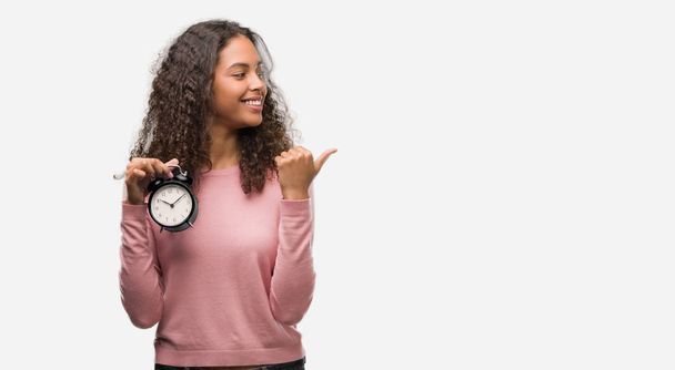 Young hispanic woman holding alarm clock pointing and showing with thumb up to the side with happy face smiling - Фото, зображення