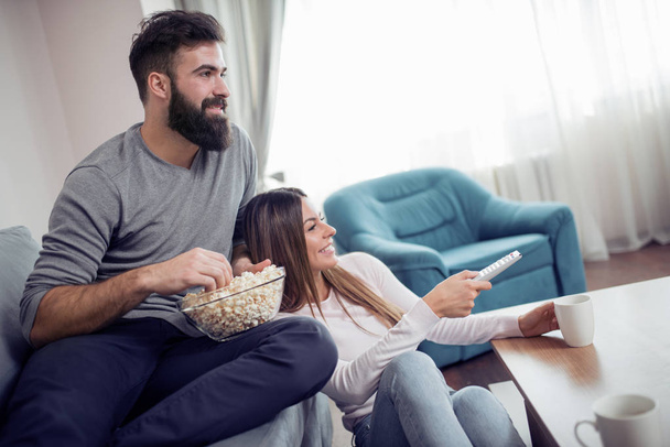 Молодая пара отдыхает в своей квартире, сидит на диване, смотрит кино и ест попкорн
.  - Фото, изображение