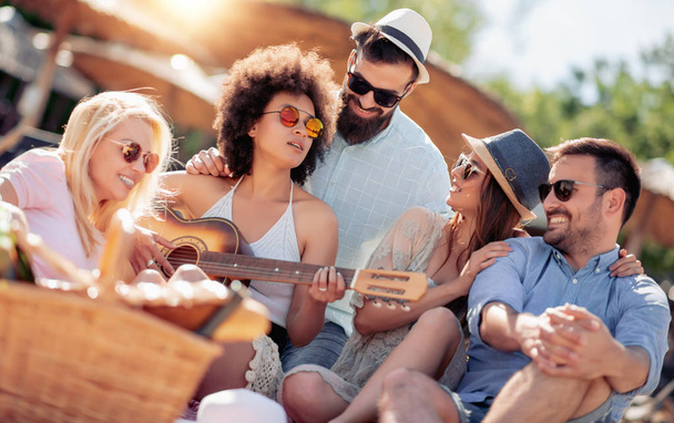 Jóvenes amigos disfrutando de una fiesta en la playa con música y bebidas.Joven tocando la guitarra con sus amigos
. - Foto, imagen