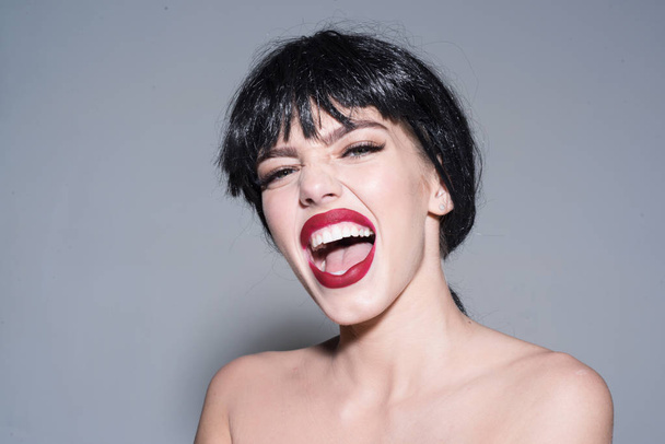Woman with attractive red lips looks at camera. Girl on scandalous shouting face posing with naked shoulders. Lady in black wig with make up on grey background. Scandalous lady concept - Photo, image