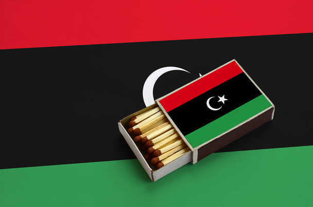 libya flag wird in einer offenen Streichholzschachtel gezeigt, die mit Streichhölzern gefüllt ist und auf einer großen Fahne liegt. - Foto, Bild