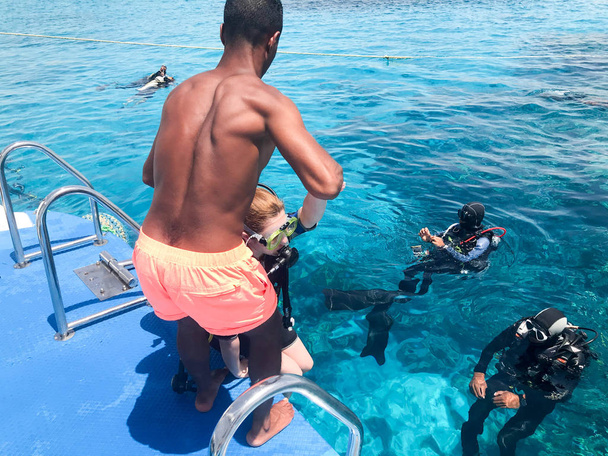 A black, swarthy, Arab athletic athletic man helping divers in black scuba diving suits with oxygen bottles sinks under the transparent blue water in the sea, the ocean in a tropical paradise warm resort. - Photo, Image