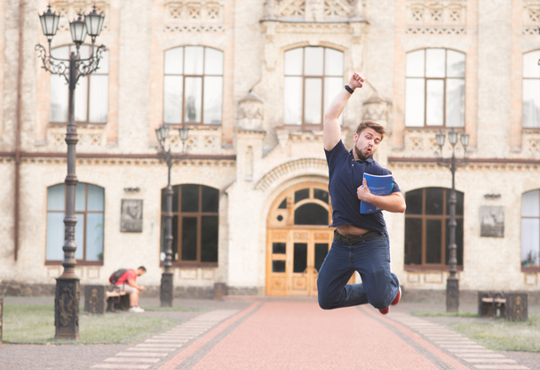 Портрет щасливого студента, який стрибає з книгами в руках на тлі університетської будівлі. Радісна людина стрибає на кампусі в університеті
. - Фото, зображення
