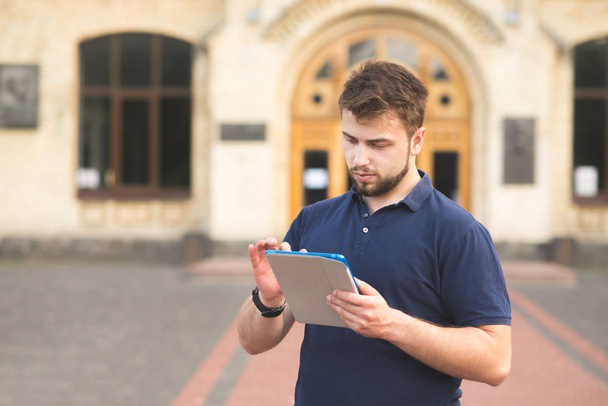Πορτρέτο ενός φοιτητή χρησιμοποιώντας ένα tablet στο φόντο ενός κτιρίου του Πανεπιστημίου. Άνδρας με μούσι στέκεται στο δρόμο με ένα tablet στα χέρια του και να χρησιμοποιεί το διαδίκτυο - Φωτογραφία, εικόνα