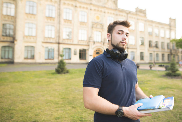 Retrato de un estudiante de pie con libros y cuadernos en el campus. Un hombre con barba y auriculares en el fondo del edificio de la universidad
. - Foto, imagen
