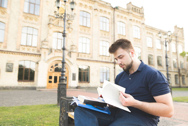 Serious student sitting on a bench at a university campus and studying. Man wearing a beard sitting on a bench on the background of a university building and reading a book. - Photo, image