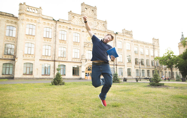 Szczęśliwy student z książek w jego rękach skacze na terenie kampusu na tle budynku Uniwersytetu. Emocjonalne człowieka z książek w jego ręce, skacze z radości. Uczeń zdał egzamin - Zdjęcie, obraz