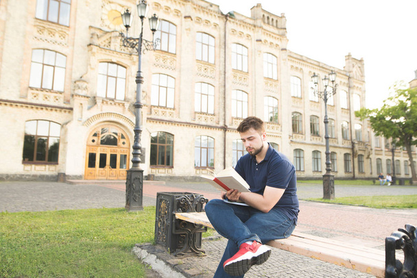 Concentrated student sitting on the background of the university and reading a book. Man with a beard sits on a bench in the campus of the university, reads a book and learns. Learning the concept - Photo, Image