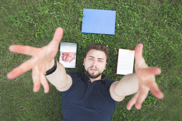 Ein gutaussehender Mann im schwarzen T-Shirt liegt auf dem Rasen neben den Büchern und streckt seine Hände in die Kamera. Porträt eines Mannes mit Bart, der im Gras liegt, Bücher am Kopf und in die Kamera blickt. - Foto, Bild
