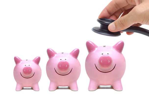 Hand holding a stethoscope over smiling pink piggy banks isolated on white   - Photo, Image