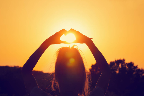 vista posteriore della donna con le mani alzate che mostrano il segno del cuore al tramonto ardente arancione
  - Foto, immagini