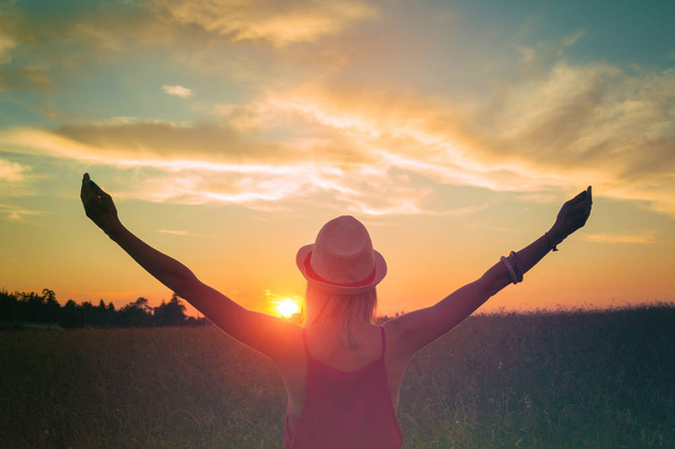 visão traseira da mulher com as mãos levantadas em pé no prado e olhando para o pôr do sol
  - Foto, Imagem