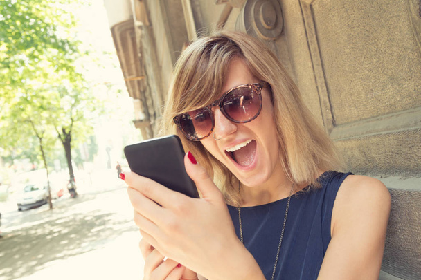 Frau mit Sonnenbrille mit Smartphone überrascht, während sie sich an Hauswand an Straße lehnt  - Foto, Bild