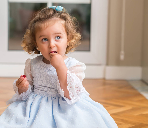 Beautiful blond child in a dress eating candy sitting on the floor at home - Fotó, kép
