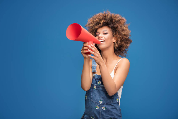 Smiling beautiful young African American woman with curly afro hair screaming by red megaphone. - Foto, imagen