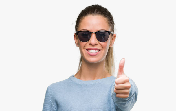 Krásná mladá žena nosí sluneční brýle a ohonu šťastný s velkým úsměvem dělá znamení ok, palec nahoru s prsty, vynikající značka - Fotografie, Obrázek