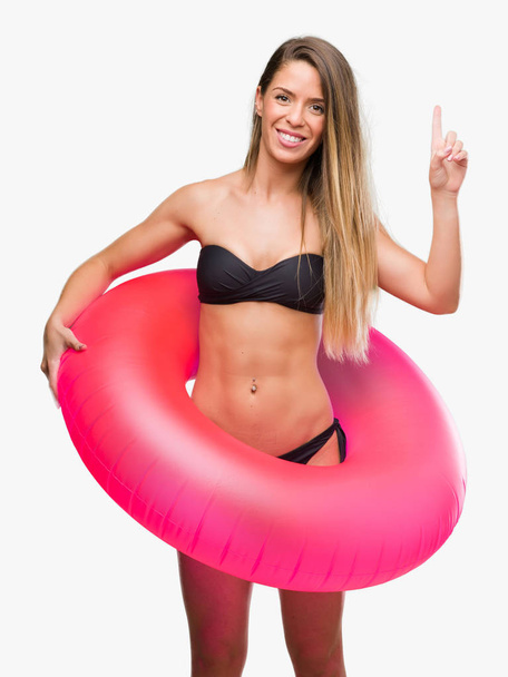 Gyönyörű fiatal nő a bikini pink gazdaság úszó meglepett mutatóujjal egy ötletet vagy kérdést boldog arc, számú - Fotó, kép