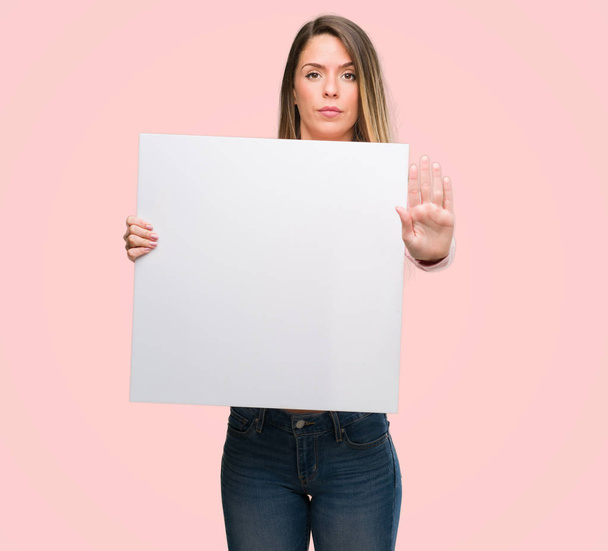 Beautiful young woman holding advertising banner with open hand doing stop sign with serious and confident expression, defense gesture - Fotó, kép