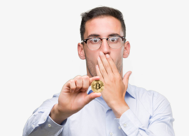 Όμορφος νεαρός άνδρας κρατά bitcoin κάλυμμα στόματος με χέρι σοκαρισμένος με ντροπή για λάθος, έκφραση του φόβου, φοβάται στη σιωπή, μυστική έννοια - Φωτογραφία, εικόνα