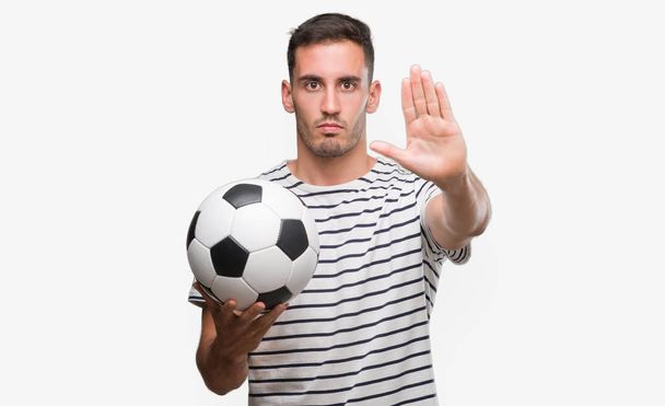 Красивый молодой человек держит футбол с открытой рукой делает знак стоп с серьезным и уверенным выражением, защитный жест
 - Фото, изображение