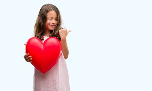 Брюнетка латиноамериканка держит красное сердце указывая и показывая с большим пальцем вверх в сторону со счастливым лицом улыбаясь
 - Фото, изображение