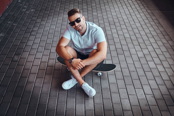 Top view portrait of a handsome fashionable skater guy in sunglasses dressed in a white shirt and shorts sitting on a skateboard on the footpath, looking at a camera. - Fotoğraf, Görsel