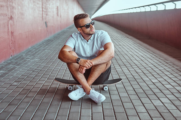 Portrait of a handsome fashionable skater guy in sunglasses dressed in a white shirt and shorts sitting on a skateboard under the bridge, looking away. - Photo, Image