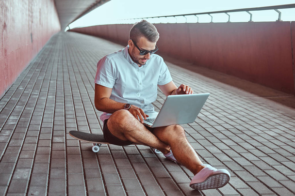 Portrait of a handsome fashionable freelancer in sunglasses dressed in a white shirt and shorts working on a laptop while sitting on a skateboard under a bridge. - Zdjęcie, obraz