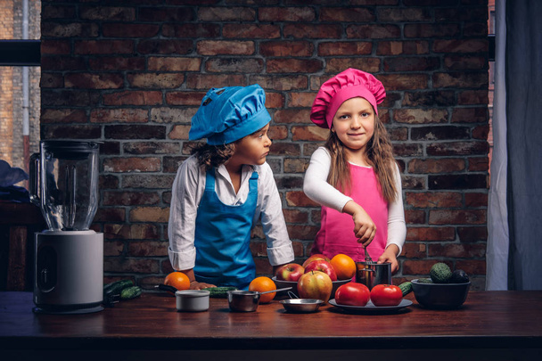 Schattig cook paar. Kleine jongen met bruin krullend haar gekleed in een blauwe cook uniform en een mooi meisje gekleed in een roze koken uniforme samen koken in een keuken tegen een bakstenen muur. - Foto, afbeelding