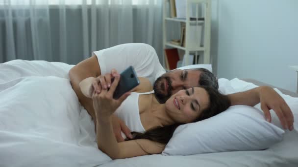 Happy couple making selfie in bed, first photo as husband and wife, family - Кадры, видео