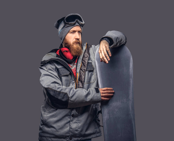 Κοκκινομάλλα βάναυση snowboarder με μια πλήρη γενειάδα στο χειμερινό καπέλο και προστατευτικά γυαλιά, ντυμένος με ένα snowboarding παλτό που ποζάρει με snowboard σε ένα στούντιο, ψάχνει μακριά. Απομονωμένοι σε ένα γκρίζο φόντο. - Φωτογραφία, εικόνα