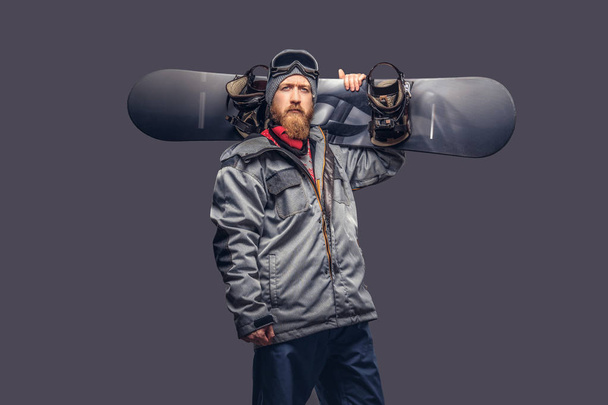Brutal redhead snowboarder with a full beard in a winter hat and protective glasses dressed in a snowboarding coat posing with a snowboard in a studio, looking at a camera. Isolated on a gray background. - Photo, Image