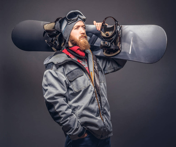 Brutal redhead snowboarder with a full beard in a winter hat and protective glasses dressed in a snowboarding coat posing with snowboard at a studio. Isolated on a gray background. - Photo, Image