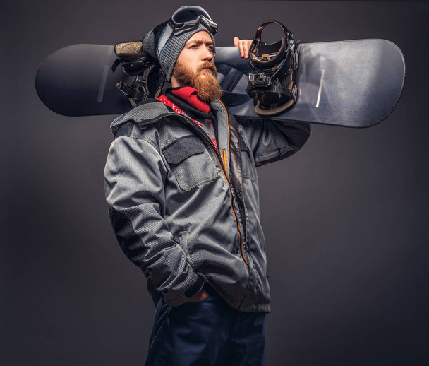 Brutaali punapää lumilautailija, jolla on täysi parta talvella hattu ja suojalasit pukeutunut lumilautailu takki poseeraa lumilauta studiossa. Eristetty harmaalla pohjalla
. - Valokuva, kuva