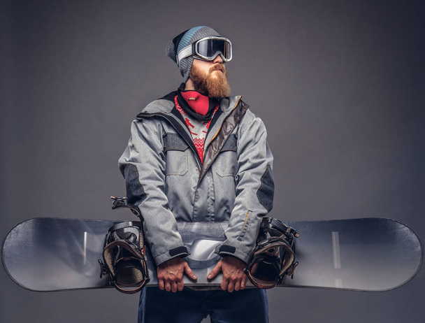 Жестокий рыжий сноубордист с бородой в зимней шляпе и защитными очками, одетый в плащ, позирующий со сноубордом в студии. Изолированный на сером фоне
. - Фото, изображение