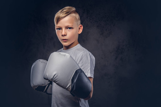 Een bokser knappe kleine jongen met blonde haren gekleed in een wit t-shirt in handschoenen klaar om te vechten. Geïsoleerd op de donkere gestructureerde achtergrond. - Foto, afbeelding