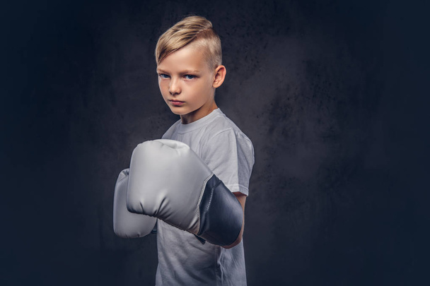 Un guapo boxeador de niño con el pelo rubio vestido con una camiseta blanca con guantes listos para pelear. Aislado sobre el fondo de textura oscura
. - Foto, imagen