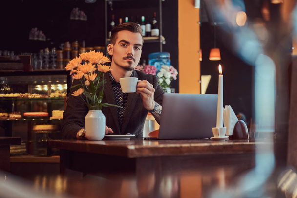 Homem freelancer bonito com barba elegante e cabelo vestido com um terno preto sentado em um café com um laptop aberto e segura uma xícara de café, olhando para a câmera
. - Foto, Imagem