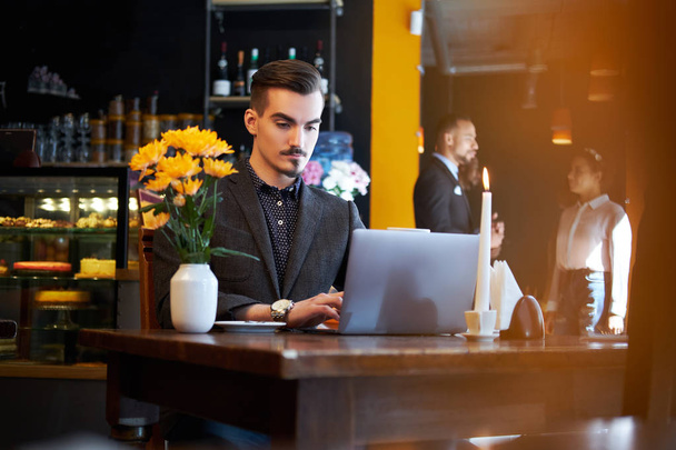 Komea freelancer mies tyylikäs parta ja hiukset pukeutunut musta puku työskentelee kannettavan tietokoneen istuessaan kahvilassa
. - Valokuva, kuva