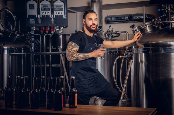 Portrait of a stylish bearded tattooed dark skinned male holds a glass of beer, standing behind the counter in the brewery - Foto, Imagem