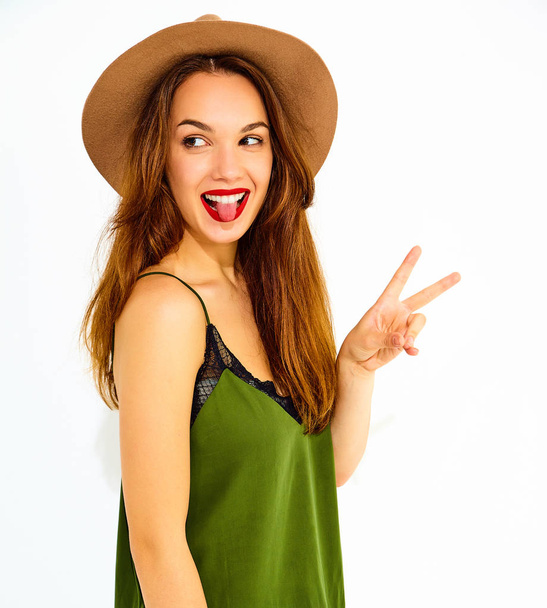 Young stylish girl model in casual summer green clothes and brown hat with red lips, showing peace sign and her tongue - Foto, Imagen