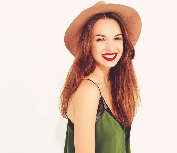 Young stylish girl model in casual summer green clothes and brown hat with red lips, posing near white wall - Fotó, kép