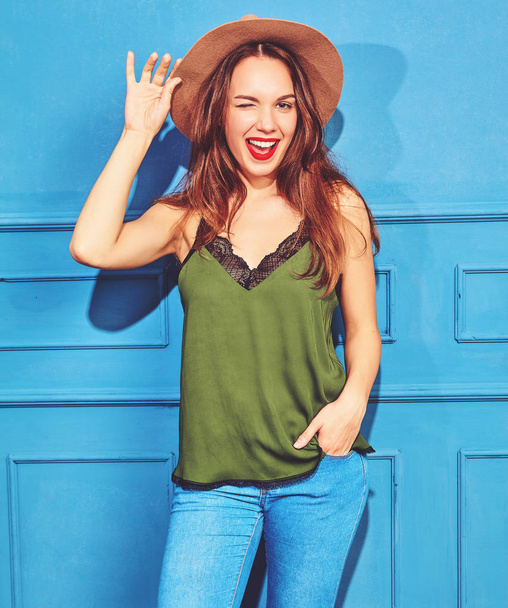 Young stylish girl model in casual summer green clothes and brown hat with red lips, posing near blue wall, winking - Foto, Imagen