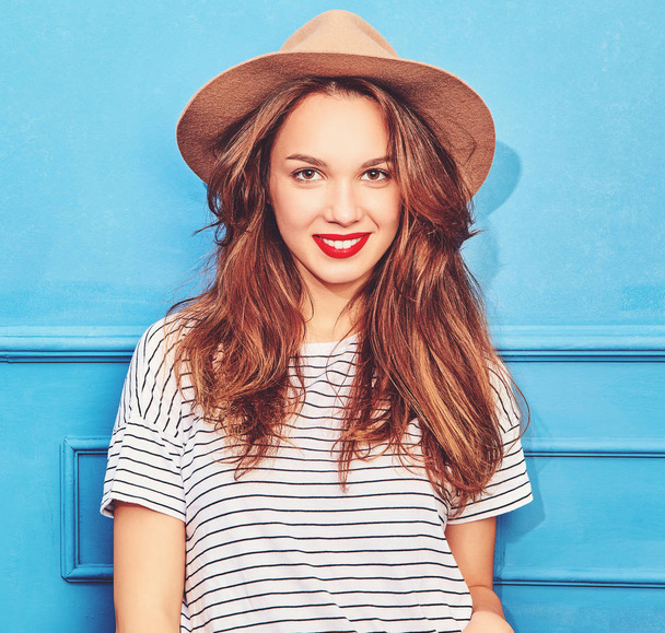 Young stylish girl model in casual summer clothes  and brown hat with red lips, posing near blue wall - Φωτογραφία, εικόνα