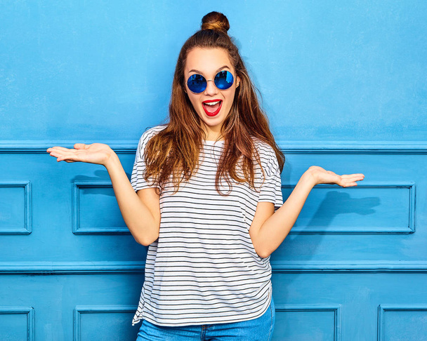 Comparison concept. Young brunette woman in casual summer hipster clothes displaying something on both flat hands for similar choice of product, posing near blue wall - Фото, зображення