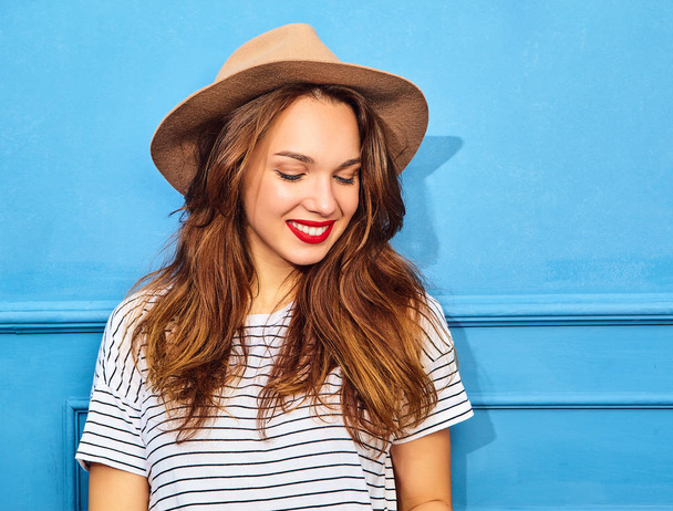 Young stylish girl model in casual summer clothes  and brown hat with red lips, posing near blue wall - Фото, зображення