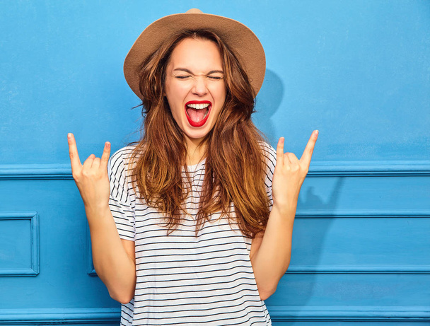 Young stylish girl model in casual summer clothes and brown hat with red lips, posing near blue wall. Showing rock and roll sign - Foto, afbeelding