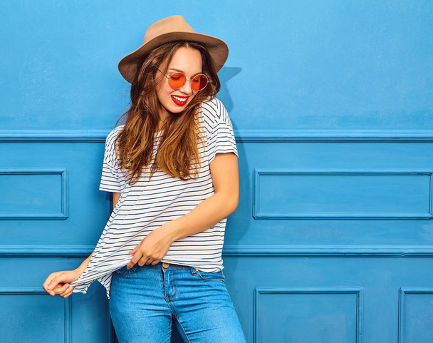 Young stylish girl model in casual summer clothes and brown hat with red lips, posing near blue wall - Foto, immagini
